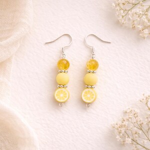 May include: A pair of dangle earrings featuring a lemon slice design. Each earring has a silver hook, a yellow bead, a row of silver beads, a round yellow bead, another row of silver beads, and a lemon slice charm.