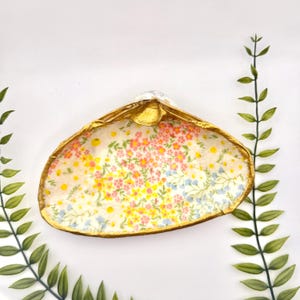 May include: A small, white ceramic dish shaped like a seashell with a gold rim. The dish is decorated with a floral pattern in shades of pink, yellow, and blue.