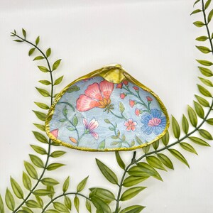 Op de afbeelding: Een decoratief schelpdier met een lichtblauw interieur met een bloemenontwerp in roze, oranje en blauw. De rand van de schelp is goudkleurig afgewerkt en de achtergrond bevat groene, bladrijke takken.