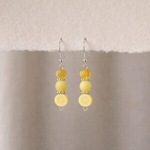 May include: A pair of dangle earrings featuring a lemon slice design. Each earring has a silver hook, a faceted yellow bead, a small silver bead, a round yellow bead, another small silver bead, and a lemon slice charm.