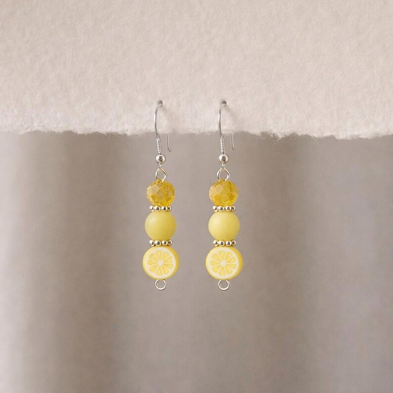 May include: A pair of dangle earrings featuring a lemon slice design. Each earring has a silver hook, a faceted yellow bead, a small silver bead, a round yellow bead, another small silver bead, and a lemon slice charm.
