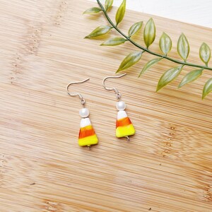 May include: A pair of silver dangle earrings with a white pearl bead and a candy corn-shaped charm. The charm is yellow, orange, and white.