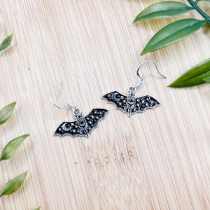 May include: A pair of silver dangle earrings featuring a black bat design with a crescent moon and stars.