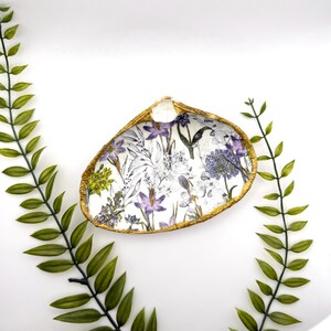 May include: A white ceramic dish shaped like a seashell with a gold rim. The dish is decorated with a floral pattern in shades of purple, white, and green.