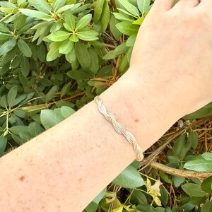 Puede incluir: Un brazalete trenzado plateado y dorado. El brazalete está hecho de hebras de metal entrelazadas, creando un patrón ondulado texturizado. La joya se exhibe sobre un fondo de follaje verde.