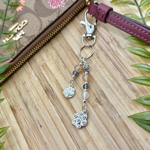 Puede incluir: Un llavero plateado con un dije de huella de pata y un adorno de cuentas. El llavero está sujeto a una correa de muñeca de cuero burdeos. El fondo es una superficie de madera con hojas verdes.