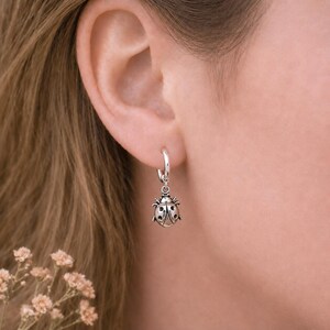Peut inclure: Une boucle d'oreille créole en argent avec une petite breloque en forme de coccinelle détaillée. Le pendentif est fabriqué en argent oxydé, mettant en valeur les ailes, les points et les pattes de l'insecte. Il s'agit d'un bijou pendant.