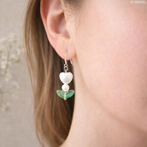 May include: Dangling earrings with a heart-shaped white pearl, a small white pearl, and a green leaf-shaped bead. The earrings are suspended from a silver hook.