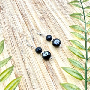 May include: A pair of black and white eight-ball earrings with silver hooks. Each earring has two black beads, with the larger bead showing the number eight in white. The earrings are on a light brown wooden surface.