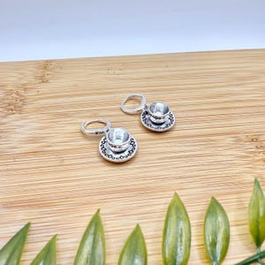 May include: A pair of silver-coloured teacup earrings. Each earring features a miniature teacup resting on a matching saucer. The teacups and saucers have intricate detailing. The earrings are displayed on a wooden surface.