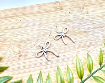 Op de afbeelding: Een paar zilverkleurige strikvormige oorbellen met haaksluitingen. De oorbellen worden gepresenteerd op een houten oppervlak, met groene bladeren op de voorgrond. De oorbellen hebben een eenvoudig, elegant ontwerp.