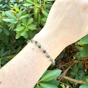 Könnte beinhalten: Ein Armband aus silbernem Draht mit kleinen, runden, dunkelgrünen Perlen. Die Perlen sind gleichmäßig auf dem Draht verteilt und bilden ein zartes Design. Das Armband wird an einem Handgelenk vor dem Hintergrund grüner Blätter gezeigt.