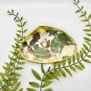 May include: A decorative seashell with a gold-painted rim and a botanical design. The interior features a collage of green leaves, brown accents, and white flowers. The shell is surrounded by green leafy branches.
