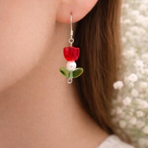 May include: A single earring featuring a red tulip design with a white pearl and green leaves. The earring is made of glass beads and has a silver hook. The earring is a floral-themed accessory.