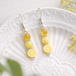 May include: A pair of dangle earrings featuring a lemon slice design. Each earring has a silver hook, yellow beads, and a lemon slice charm. The earrings are on a white, decorative plate.