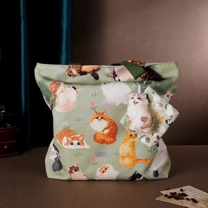 Peut inclure: Un sac cabas en toile vert avec un motif imprimé de chats. Le sac a une bandoulière verte et une petite pochette assortie avec un porte-clés chat.