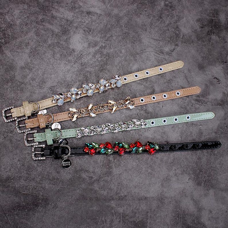 May include: Five different dog collars with various embellishments. The collars are made of leather and feature rhinestones, crystals, and beads. The collars are in different colors, including black, brown, and green.