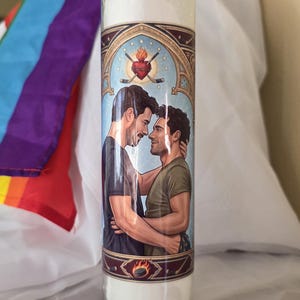 May include: A white candle in a glass jar with a decorative label featuring two men embracing. The label has a blue background with a heart and hockey sticks. A rainbow flag is visible in the background.