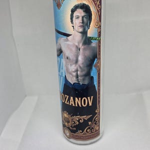 May include: A clear glass candle with a decorative label featuring a man holding a hockey stick. The label has a blue background with the name "OZANOV" in gold lettering. The candle has a religious theme.