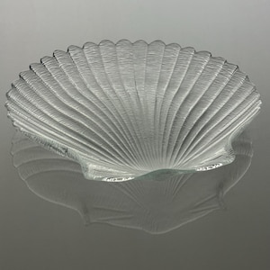 May include: A clear glass dish shaped like a scallop shell. The dish features a textured surface with radiating ridges that extend from the base to the scalloped edge, creating a translucent appearance with a reflective quality on the surface.