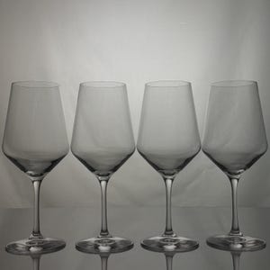 May include: A set of four clear glass wine glasses arranged in a row. Each glass features a wide, angular bowl that tapers toward the rim, supported by a slender stem and a circular base. The glassware is transparent with a smooth finish.