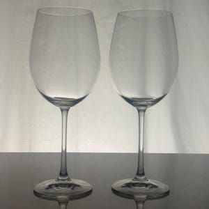 May include: Two clear glass wine glasses with tall, slender stems and wide, circular bases. Each glass features a large, rounded bowl that tapers toward the rim, designed for holding liquid. The glasses are transparent and made of smooth glass.