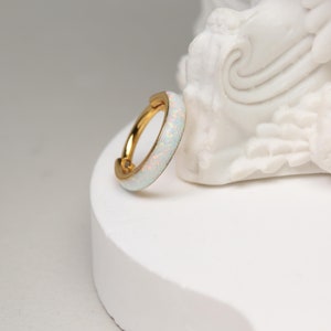 May include: A gold hoop earring with a white opal inlay. The earring is on a white pedestal.