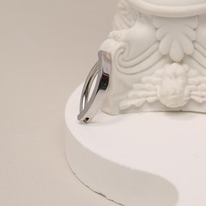 May include: A silver-colored ring with a curved design. The ring is made of a shiny metal and has a smooth surface. The ring is displayed on a white, sculpted base.