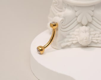 Puede incluir: Joyería corporal dorada con piedras preciosas transparentes. La joyería es curva y tiene una pequeña bola en cada extremo.