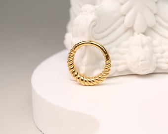 May include: A gold-colored, hinged hoop earring with a twisted rope design. The earring is circular and has a clasp closure. The jewelry is displayed on a white surface with a white sculpture in the background.