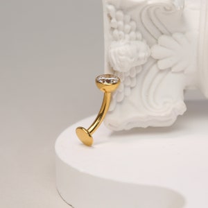 May include: Gold body jewellery with a clear gem. The jewellery is curved and has a small ball on the end.