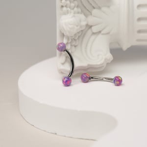 Pode incluir: Dois piercings de corpo curvos de aço cirúrgico com bolas de opala. As bolas são uma mistura de rosa, roxo e amarelo.