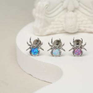 May include: Three silver spider earrings with opal stones. The stones are blue, white, and pink.