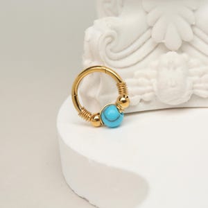 Puede incluir: Pendiente de aro dorado con una perla turquesa y detalles dorados.