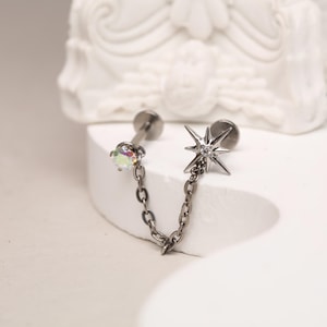 May include: A silver chain connects two body jewelry pieces. One piece features a clear iridescent gem, the other a star with a clear gem in the center.