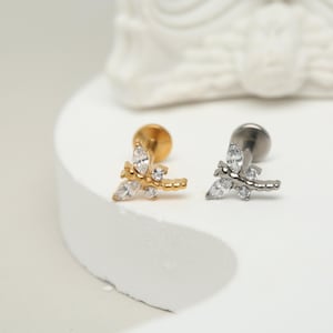 Pode incluir: Dois brincos de botão de libélula prateados e dourados com detalhes de cristal transparente. Os brincos estão sobre uma superfície branca.