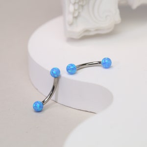 May include: Two curved eyebrow rings with blue opal-like spheres at each end. The jewelry is made of silver-colored metal. The curved shape is designed to fit the natural curve of the eyebrow. The background is white.