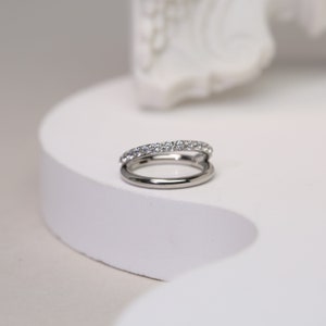 Puede incluir: Un anillo de plata con una fila de pequeñas piedras transparentes alrededor de la banda. El anillo está abierto y tiene una segunda banda que pasa por la abertura.