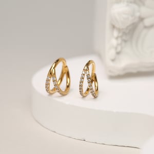 May include: Two gold-colored hoop earrings with a double-layered design. Each hoop is adorned with small, clear gemstones. The earrings are displayed on a white surface, showcasing their intricate details and elegant style. The jewelry is designed for pierced ears.