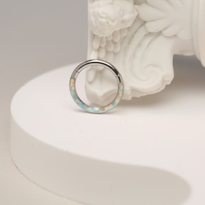 Puede incluir: Un anillo de plata con una incrustación de ópalo azul claro y blanco. El anillo está sobre una superficie blanca.