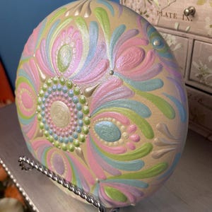 May include: A pastel-colored, hand-painted decorative stone with a floral design. The stone features a beige base with swirling patterns in pink, blue, green, and white. The stone is displayed on a silver metal stand, showcasing the intricate details of the artwork.