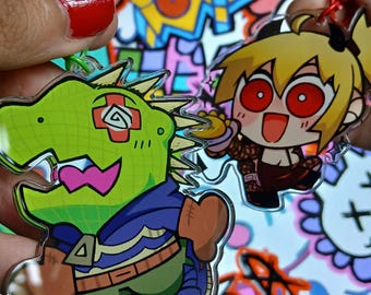 Può includere: Due portachiavi di personaggi dei cartoni animati. Uno è una creatura verde simile a un dinosauro con un abito blu e verde. L'altro è una ragazza bionda con gli occhi rossi. Entrambi i portachiavi sono tenuti da una persona con lo smalto rosso.