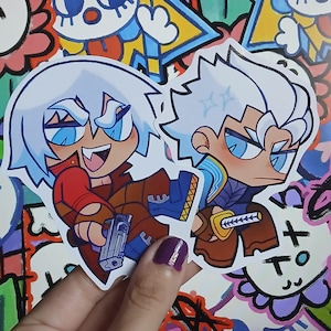 Puede incluir: Una pegatina en forma de corazón con dos personajes de estilo anime. Uno tiene el pelo blanco, una chaqueta roja y pantalones marrones, sosteniendo un arma. El otro tiene el pelo blanco, un atuendo azul y marrón y una espada.