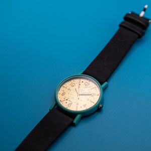 May include: A wristwatch featuring a round teal case and a black strap. The watch face is gold with black speckles and black numerals at the 1, 2, 4, 5, 7, 8, 10, and 11 o'clock positions, with a larger black numeral 12 at the top.
