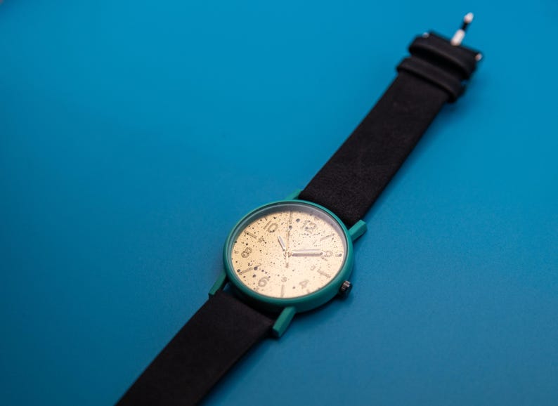 May include: A wristwatch featuring a round teal case and a black strap. The watch face is gold with black speckles and black numerals at the 1, 2, 4, 5, 7, 8, 10, and 11 o'clock positions, with a larger black numeral 12 at the top.