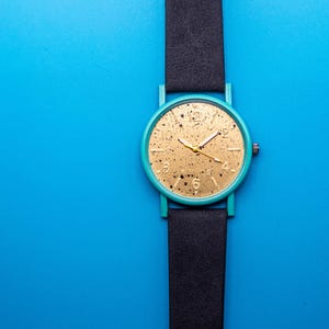 May include: A wristwatch featuring a round teal case and a black strap. The watch face is gold with a black speckled pattern, gold-tone hour markers, and gold hands. A small crown is positioned on the right side of the circular watch case.