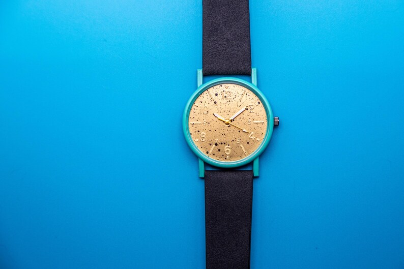 May include: A wristwatch featuring a round teal case and a black strap. The watch face is gold with a black speckled pattern, gold-tone hour markers, and gold hands. A small crown is positioned on the right side of the circular watch case.