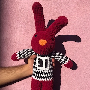 Puede incluir: Un conejo de crochet rojo con una camisa a cuadros blanco y negro con un número blanco "11" en la parte delantera.