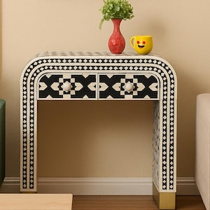 May include: A decorative console table with a black and white geometric pattern. The table has two drawers and gold-colored feet. A red vase with greenery and a yellow smiley face mug sit on top. A painting of trees hangs above.