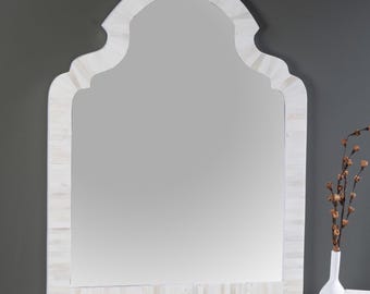 May include: A large, arched mirror with a white, mosaic-style frame. The mirror has a decorative top edge and is set against a dark gray wall. A white vase with dried stems sits to the side, adding a touch of elegance.